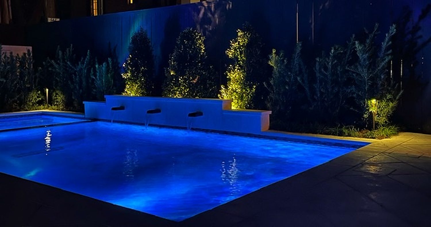 A pool lit blue at night, three sheer water features and an uplit screen of trees and palms