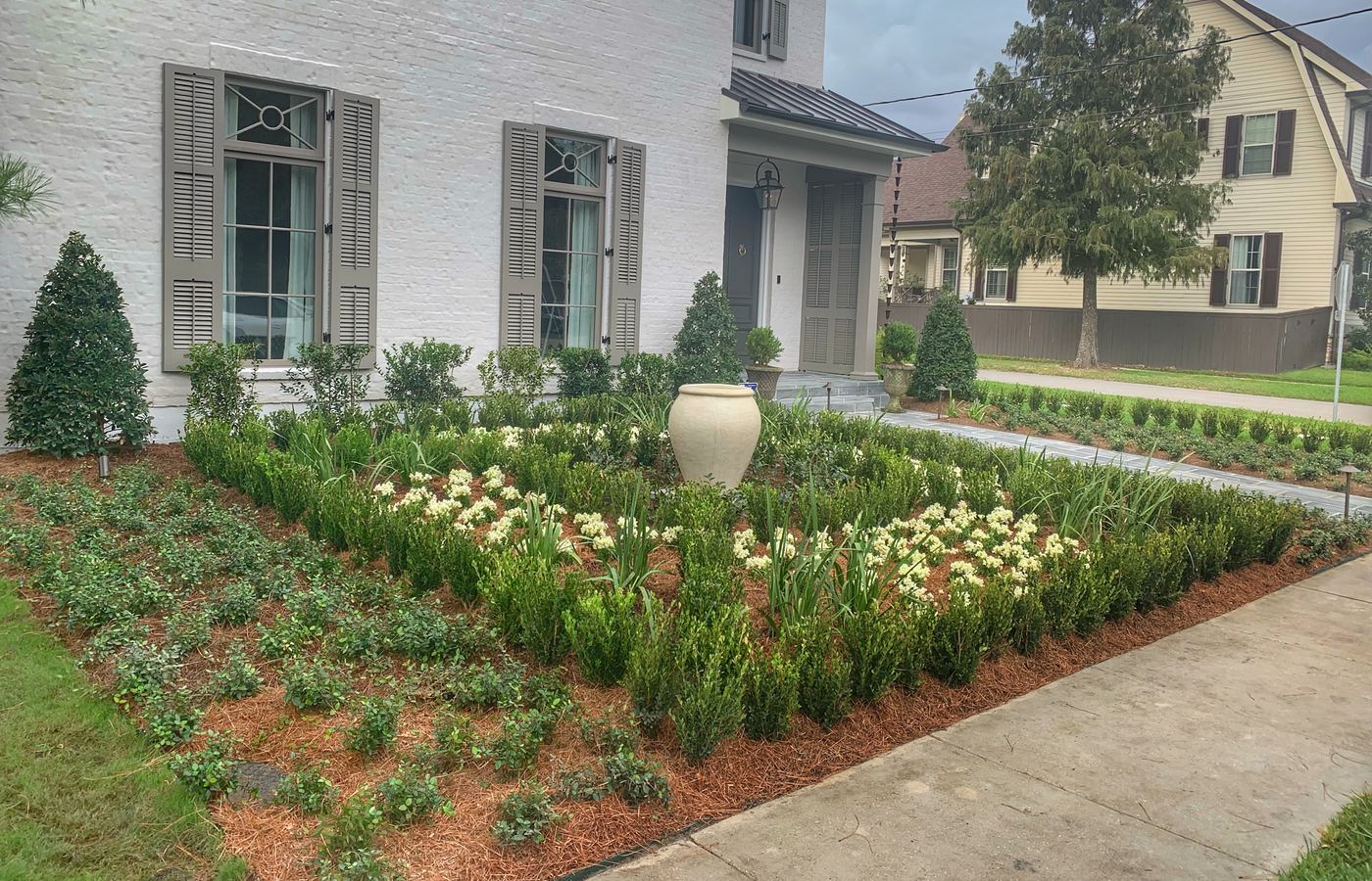 A formal parterre front garden with boxwood-edged beds, white annuals and an urn at the center