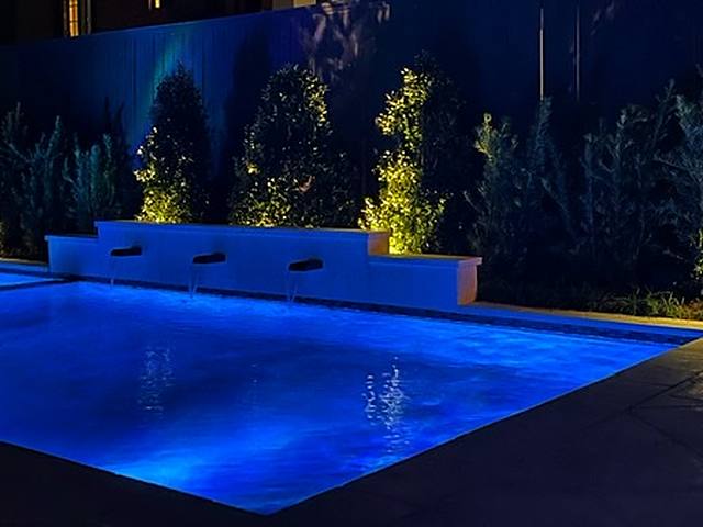 A pool lit blue at night, three sheer water features and an uplit screen of trees